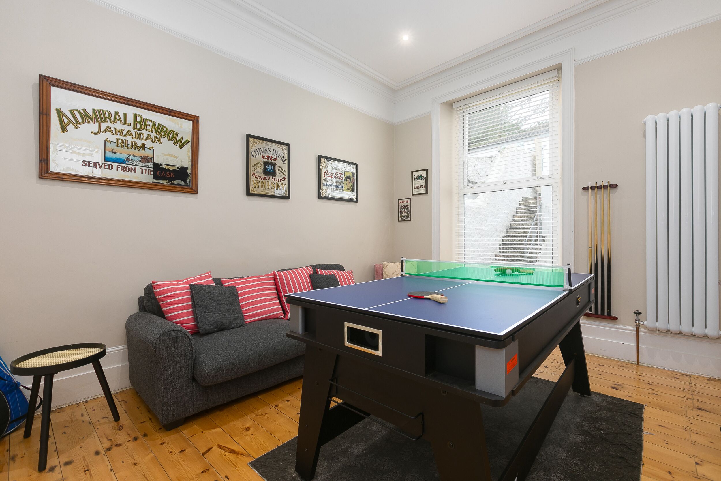 Une pièce avec une table de billard, un canapé gris orné de coussins à rayures rouges et blanches, une petite table ronde et un panneau encadré accroché au mur.