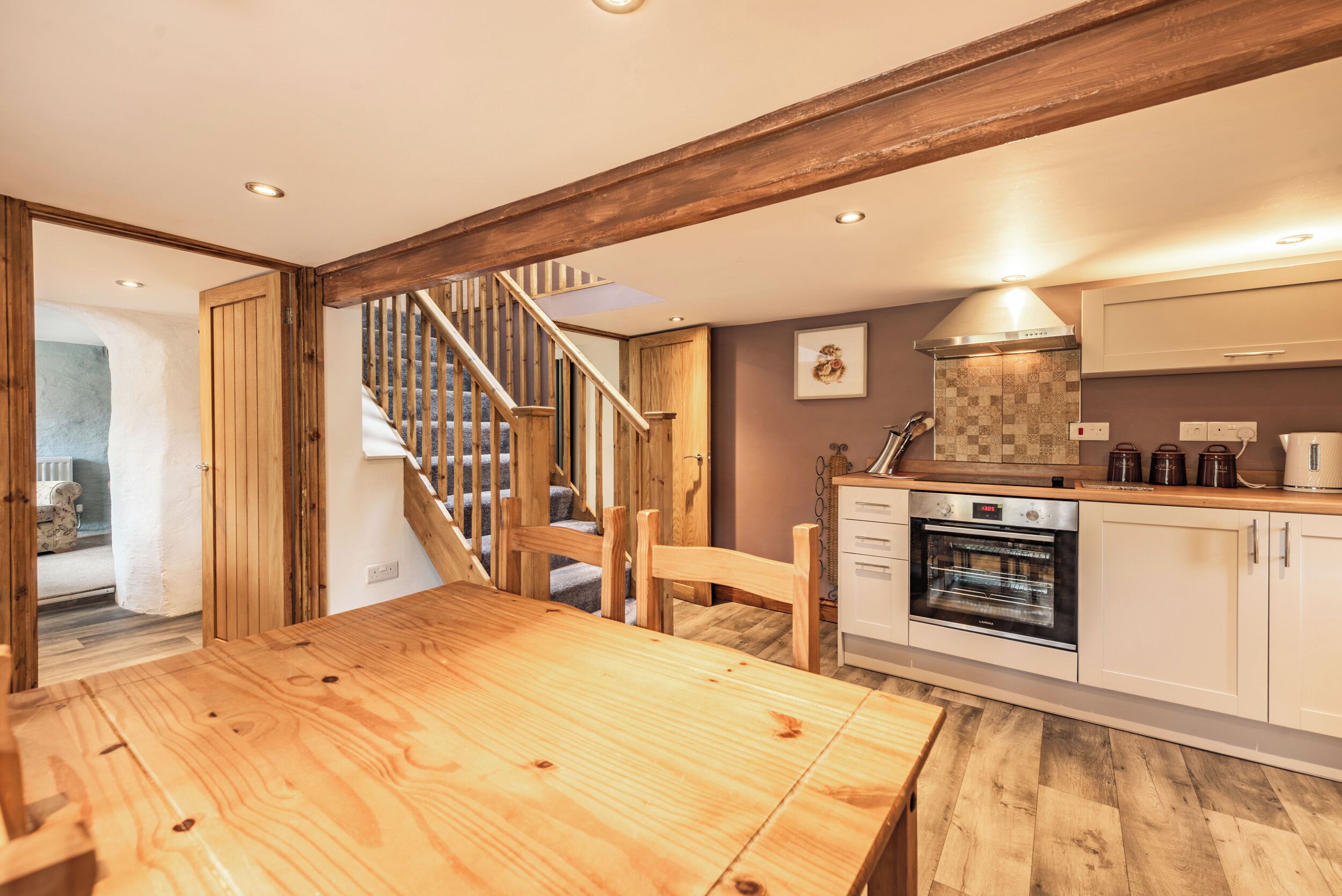 A wooden dining table with chairs, a kitchen with white cabinets, a built-in oven, and a staircase with wooden railings.