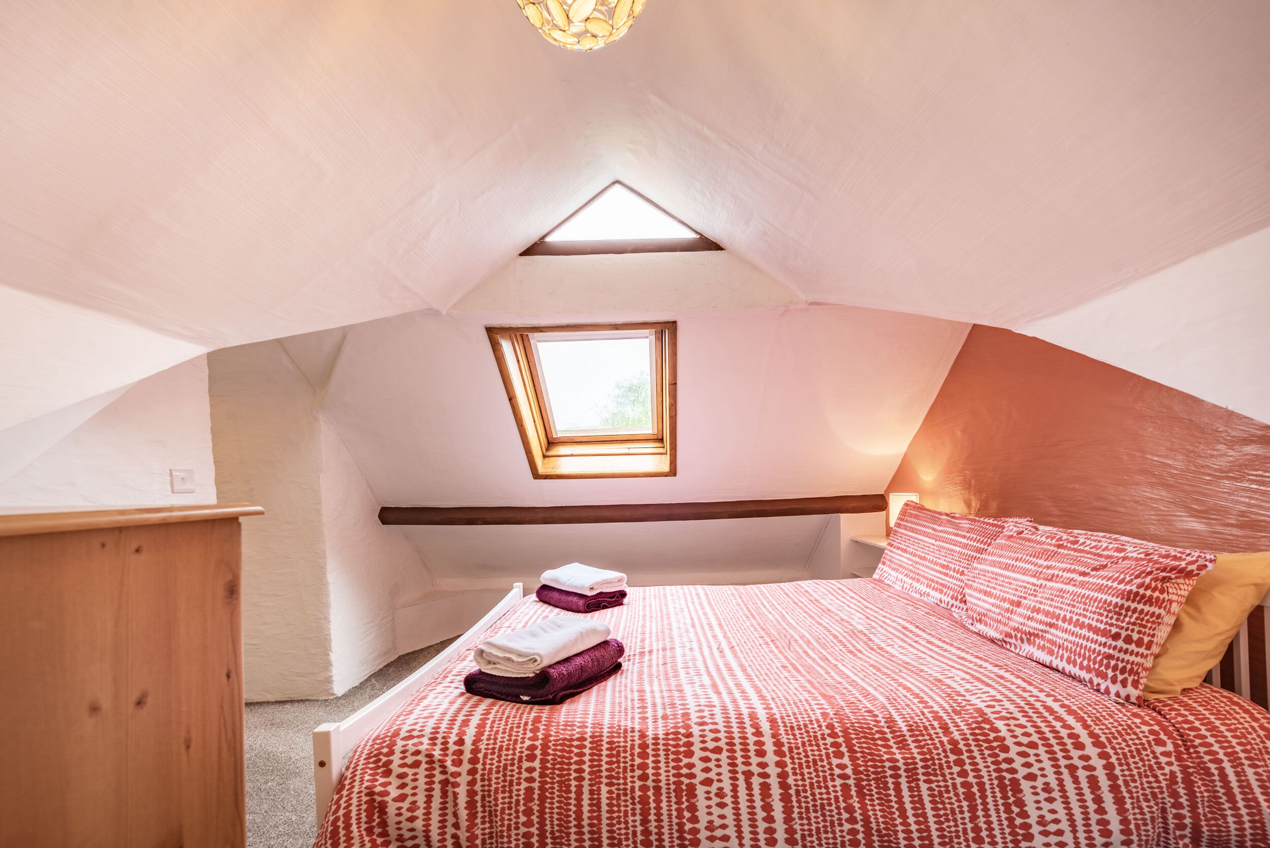 A cozy attic bedroom with a sloped ceiling, a skylight, a neatly made bed with a patterned bedspread, and a small bedside table.