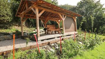 A wooden gazebo with a bench, surrounded by a garden with gravel paths and plants.