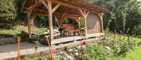A wooden gazebo with a bench, surrounded by a garden with gravel paths and plants.