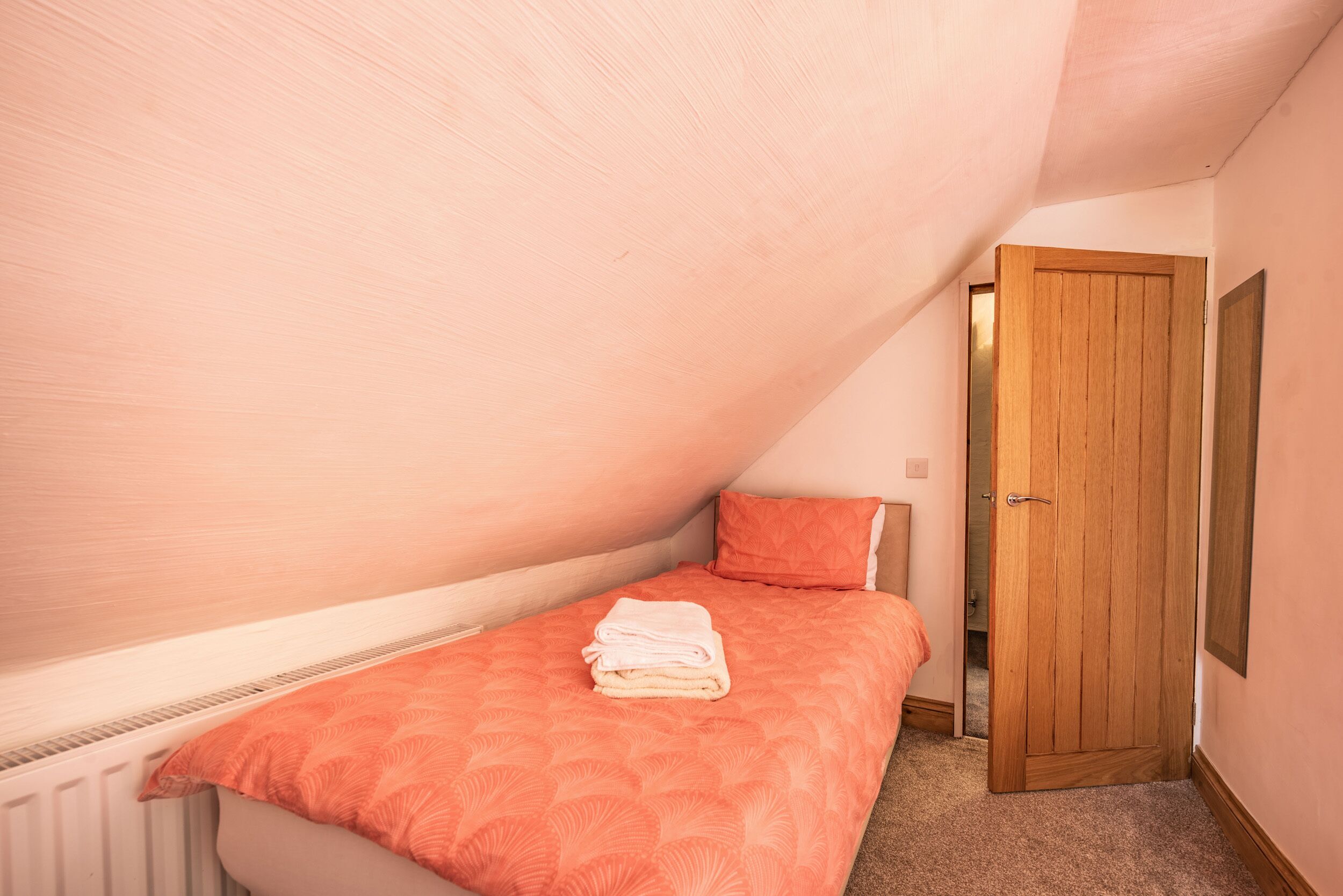 A bedroom with a sloped ceiling, a bed with an orange bedspread, a wooden door, and a radiator.