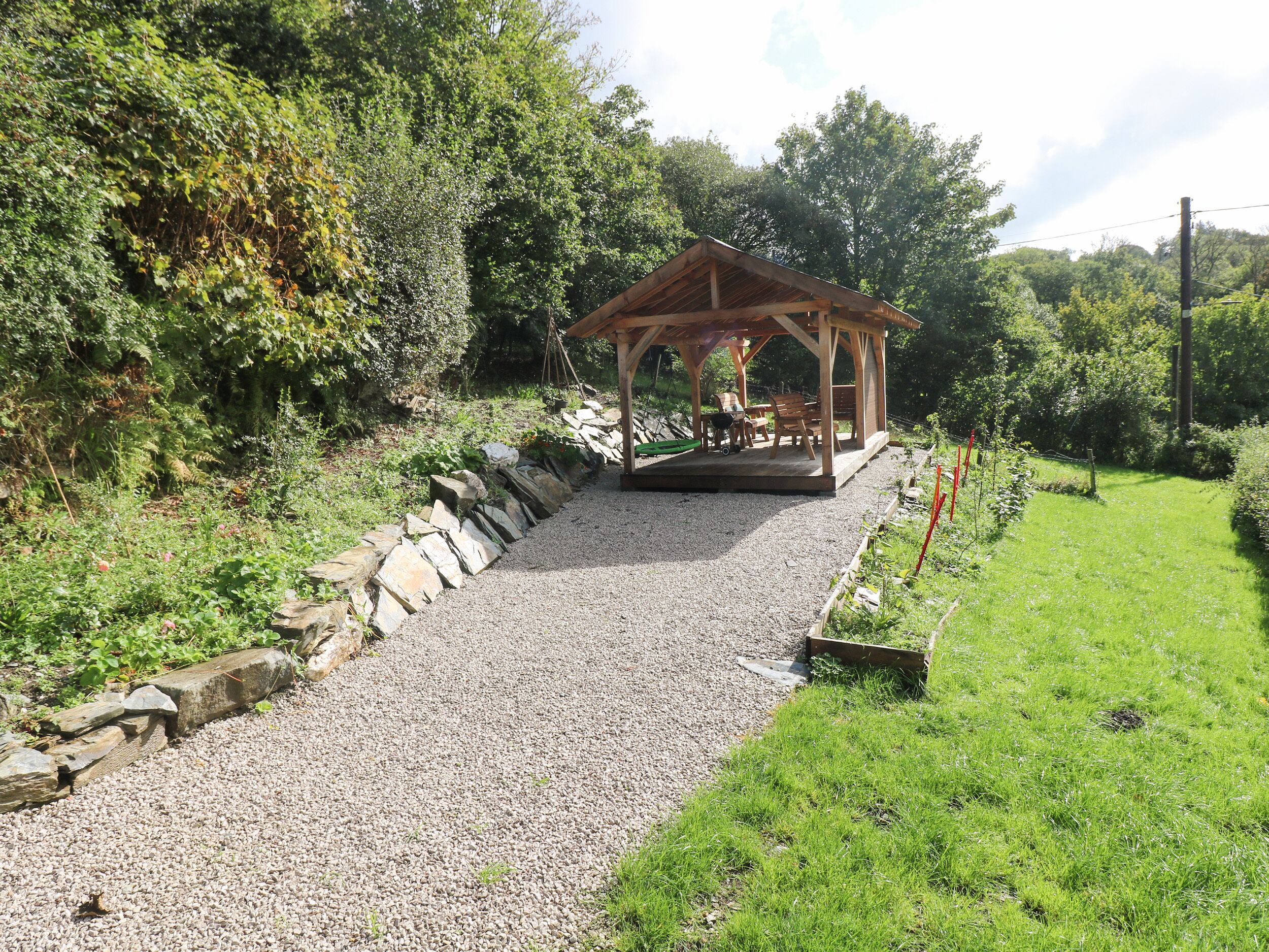 A wooden gazebo with a gravel path leading to it, surrounded by greenery and a stone wall.