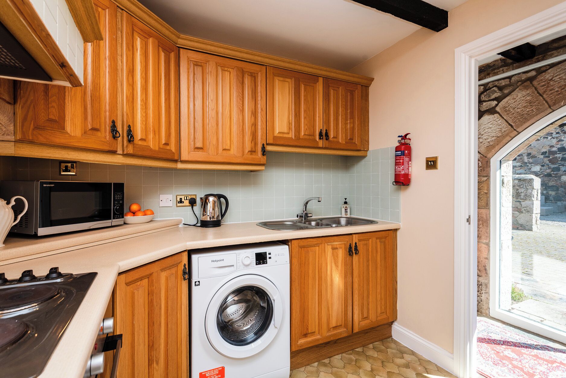 A kitchen with wooden cabinets, a microwave, a kettle, a sink, and a washing machine.