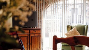 A room with a television on a wooden cabinet, a green armchair with a cushion, and a large window with vertical blinds. - Lupin Cottage (Wooler)
