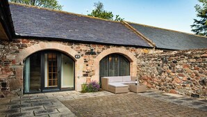 A stone building with two arched windows and a stone wall. - Lupin Cottage (Wooler)
