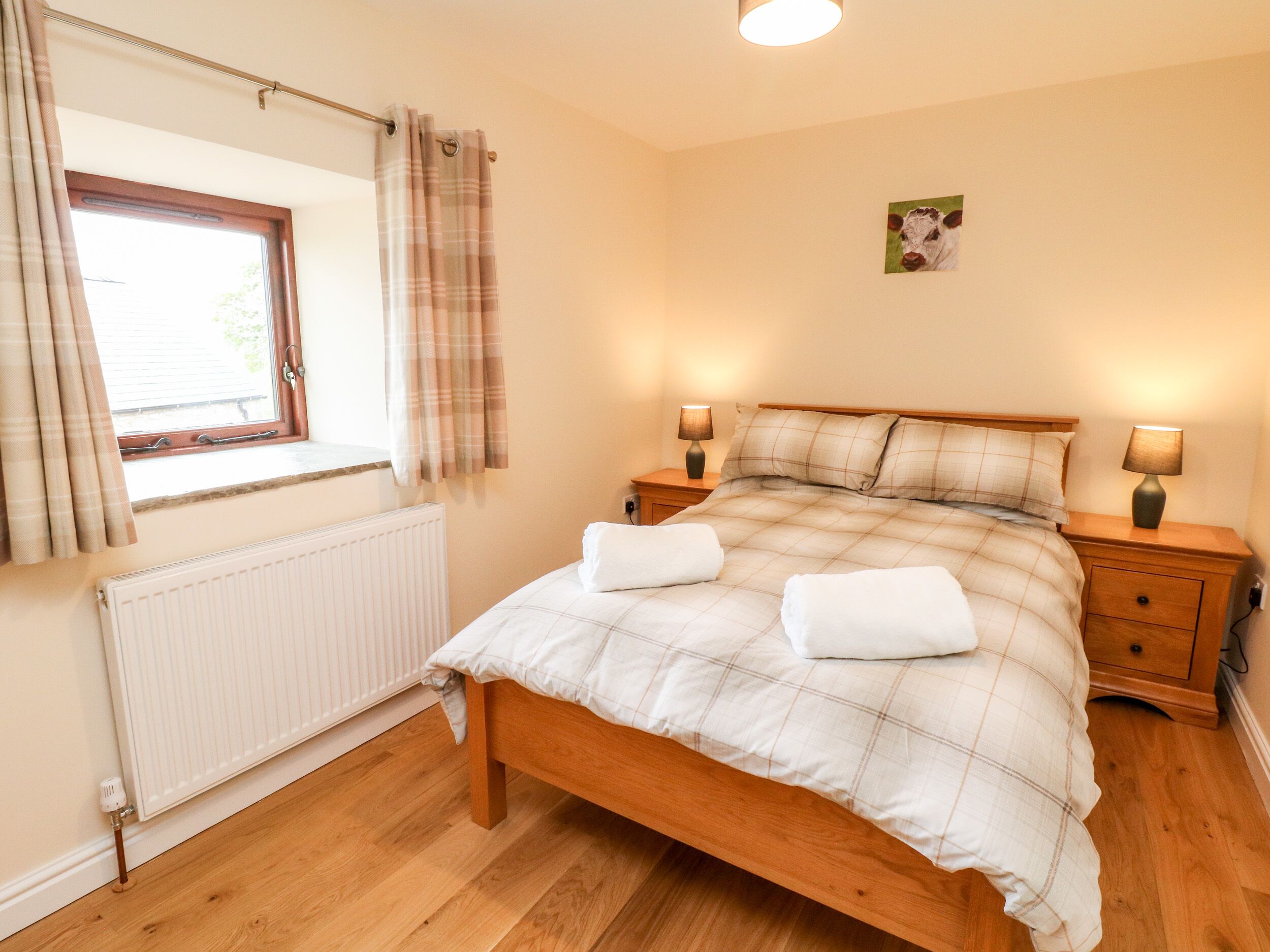 A neatly made bed with a plaid bedspread, two bedside lamps, a radiator, and a window with curtains.