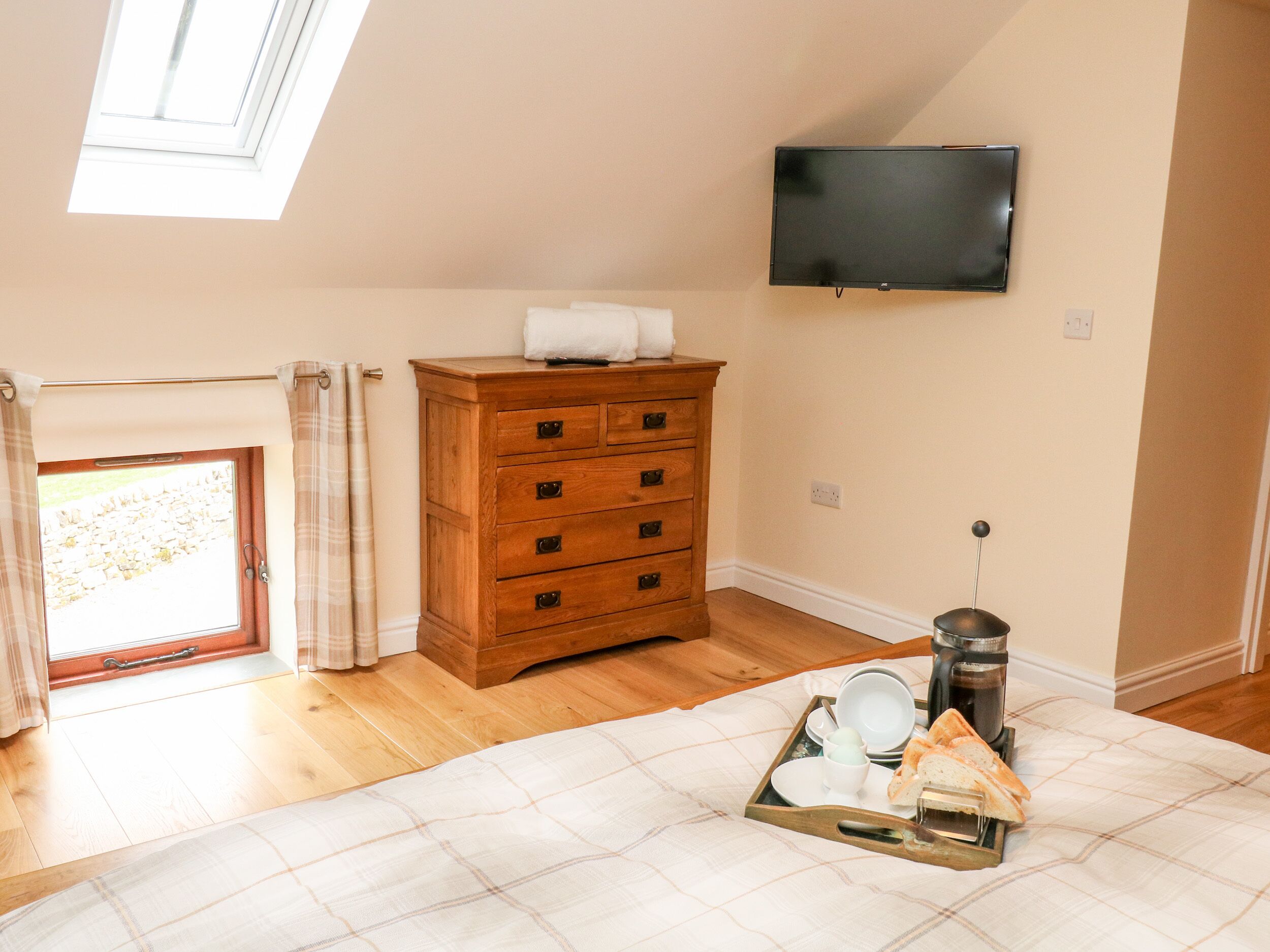 A bedroom with a wooden dresser, a flat-screen TV mounted on the wall, a window with curtains, and a breakfast tray on the bed.