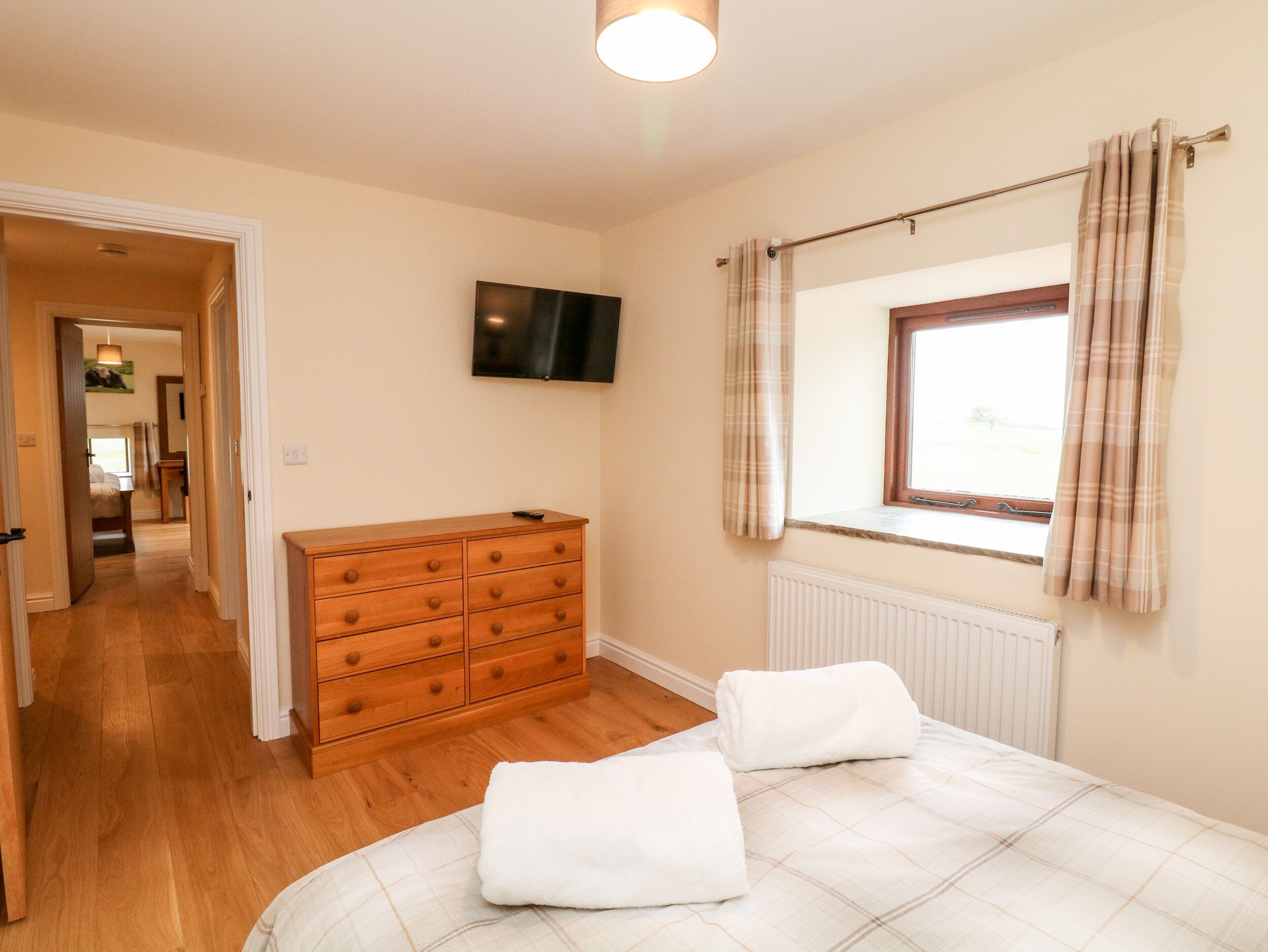 A bedroom with a wooden dresser, a bed with white bedding, a television on the wall, and a window with curtains.