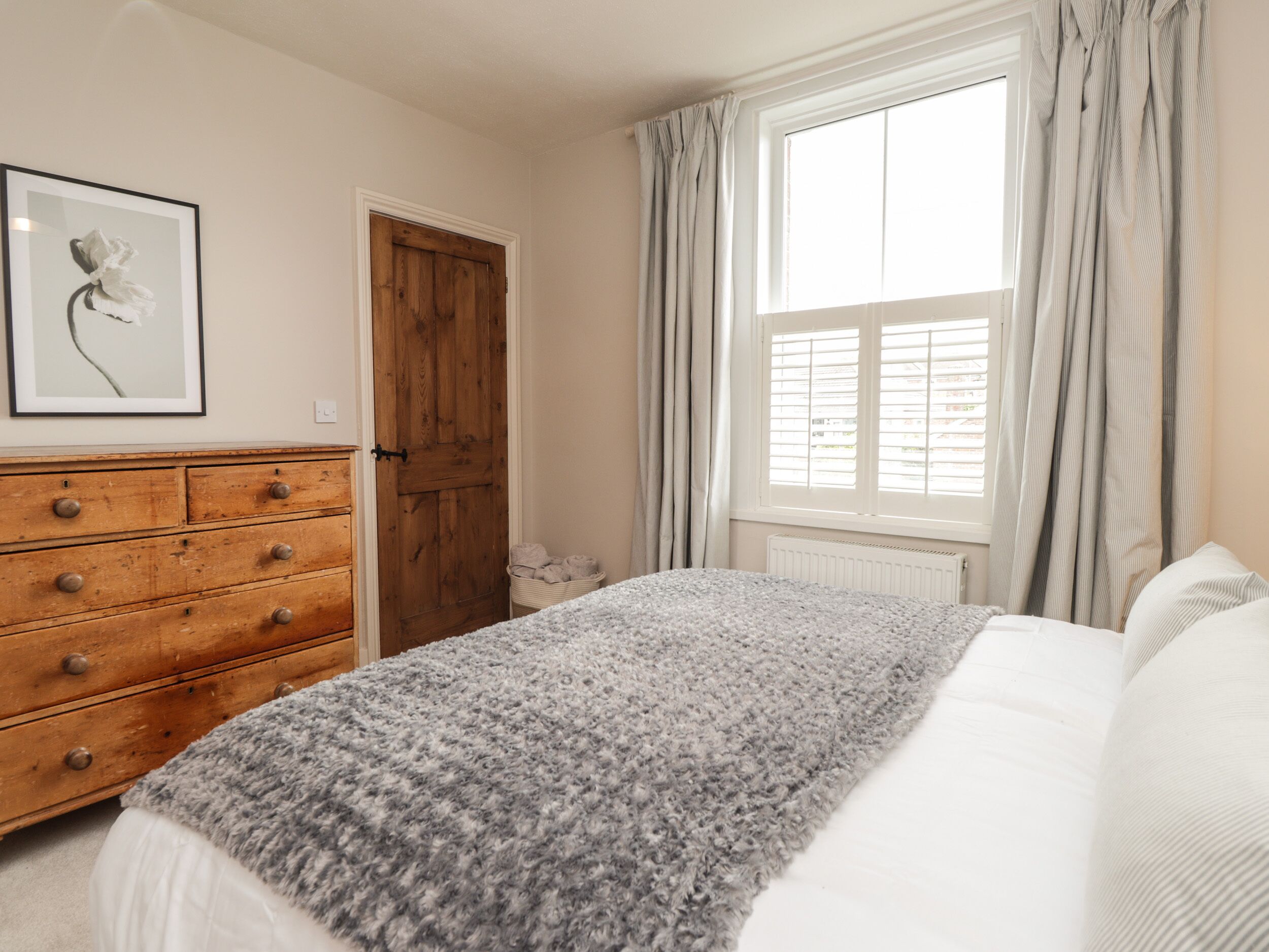 A bedroom with a wooden dresser, a bed with a grey blanket, a window with curtains, and a door.