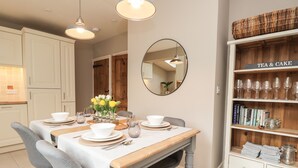 Un comedor con una mesa de madera dispuesta para cuatro personas, rodeada de sillas grises. Un espejo cuelga de la pared sobre la mesa, y a la derecha hay una estantería llena de libros y cristalería.