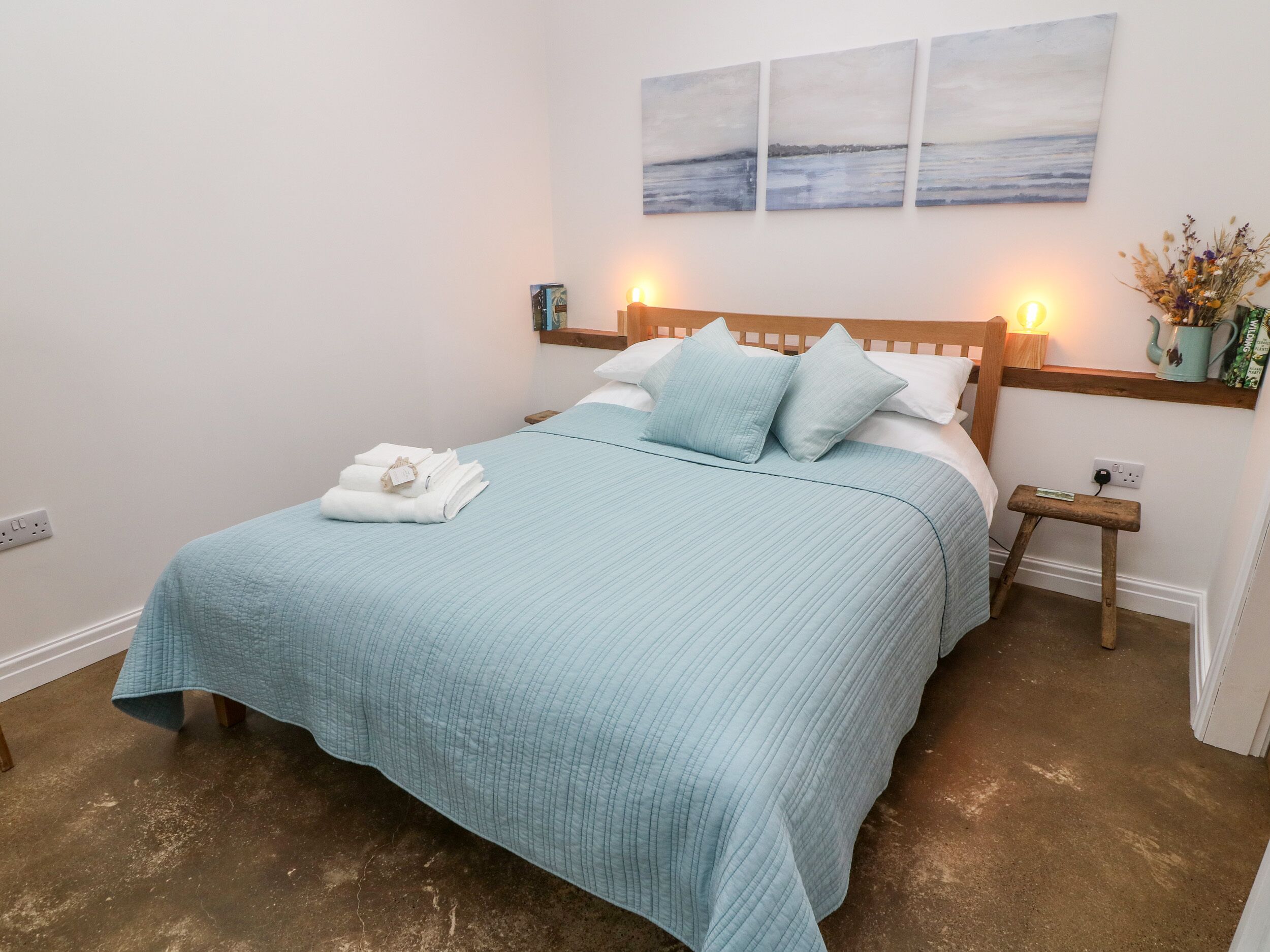 A neatly made bed with a blue quilt, two pillows, and folded towels on a wooden headboard. There are wall-mounted paintings above the bed, and a small table with a vase of flowers to the right.
