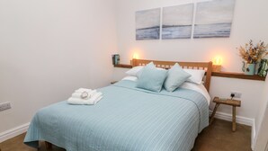 A neatly made bed with a blue quilt, two pillows, and folded towels on a wooden headboard. There are wall-mounted paintings above the bed, and a small table with a vase of flowers to the right. - Horncop Nook (Kendal)