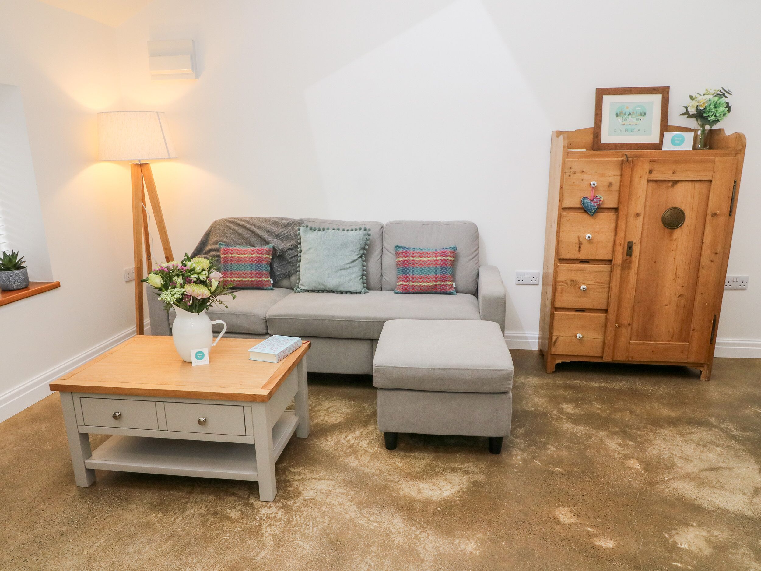 Ein Wohnzimmer mit grauem Sofa, einem Holzschrank, einem kleinen Tisch mit Blumenvase und einer Stehlampe.