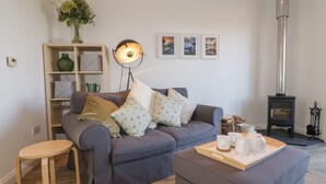 A living room with a grey sofa, a small wooden table, a floor lamp, a bookshelf, and a wood-burning stove. - Badgers Way (Tintagel)