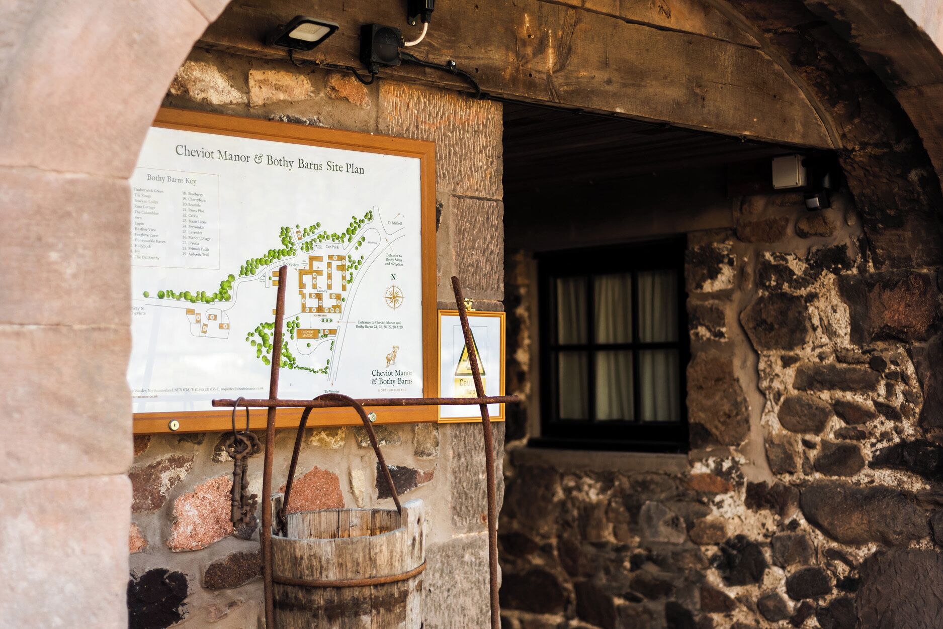 Een stenen gebouw met een poster van een Cheviot Manor & Bothy Barns plattegrond, een houten emmer en een raam.