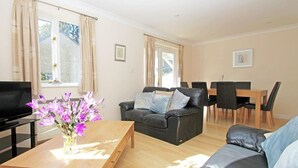 Ein Wohnzimmer mit einer Couch, einem Couchtisch mit Blumenvase, einem Fernseher und einem Essbereich mit Stühlen und Tisch.