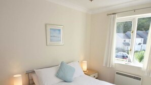 Ein ordentlich bezogenes Bett mit weißer Bettwäsche und einem blauen Kissen, flankiert von zwei Nachttischen mit Lampen, einem gerahmten Bild an der Wand und einem Fenster mit Blick auf Häuser und ein Auto draußen.