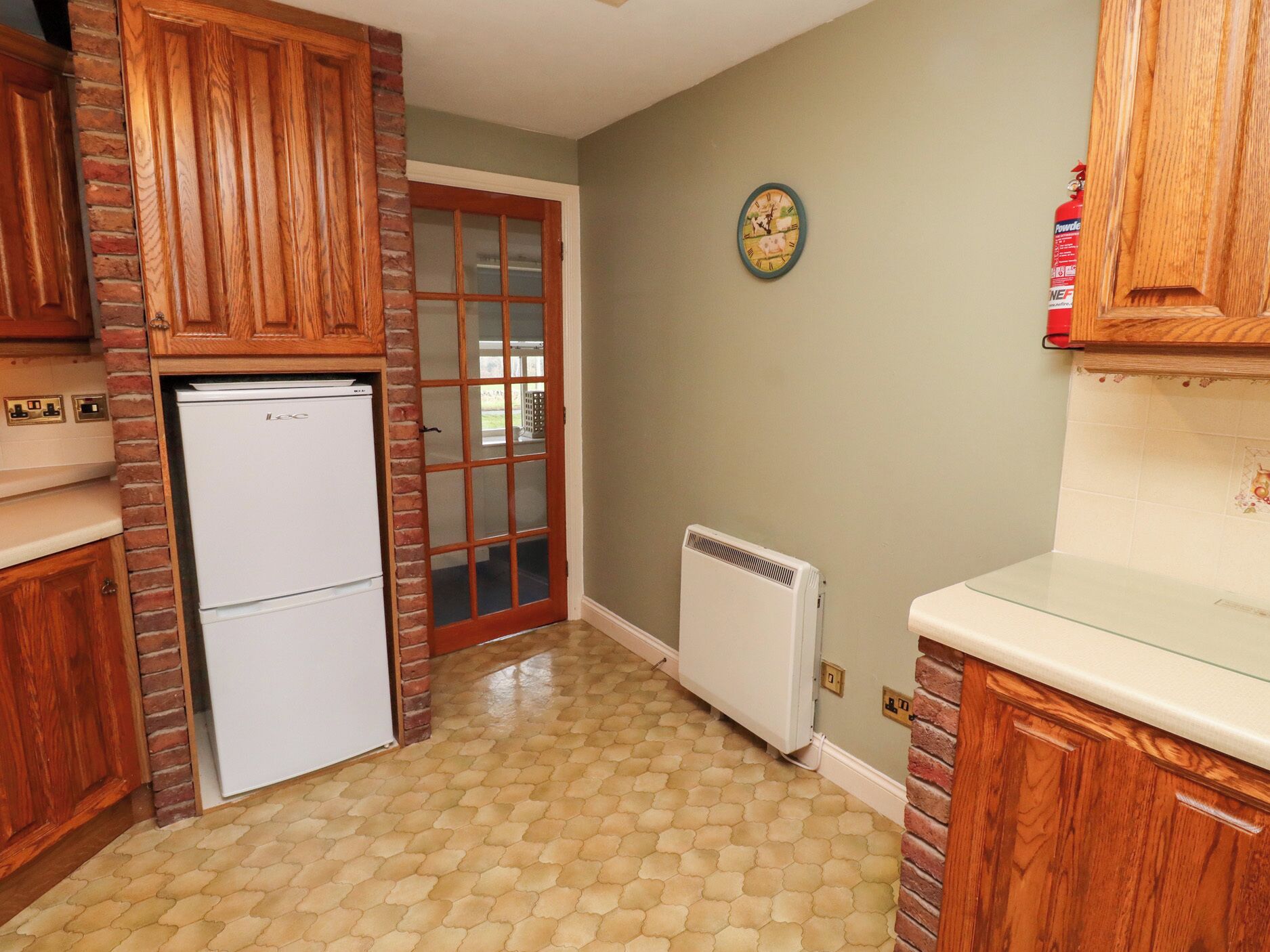 Une cuisine avec des meubles de rangement en bois, un réfrigérateur blanc, une porte en verre, une horloge murale et un radiateur blanc.