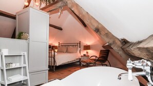 Un dormitorio con una bañera blanca grande, techo de madera, cama con cabecera, mesita de noche y una silla.