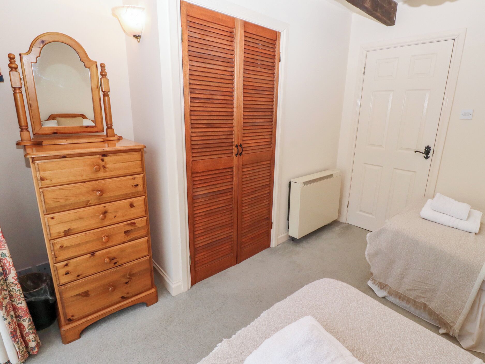 A bedroom with a wooden dresser, a large mirror, a wardrobe, a radiator, and a bed with a white blanket.