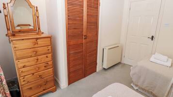 Une chambre avec une commode en bois, un grand miroir, une armoire, un radiateur et un lit recouvert d’une couverture blanche.