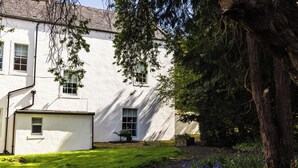 A white building with multiple windows, a small wheelbarrow, and a tree in the foreground. - Pansy Plot (Wooler)