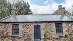 A stone building with a dark blue door, two windows with shutters, and a stone chimney. - Bracken Lodge (Wooler)