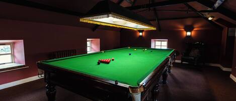 A pool table with balls, a chair, and a window in a dimly lit room with a high ceiling.