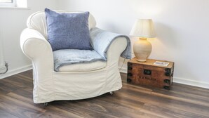 Ein weißer Sessel mit blauer Kissenauflage und einer Decke, ein Holznachttisch mit Lampe sowie ein alter Koffer.