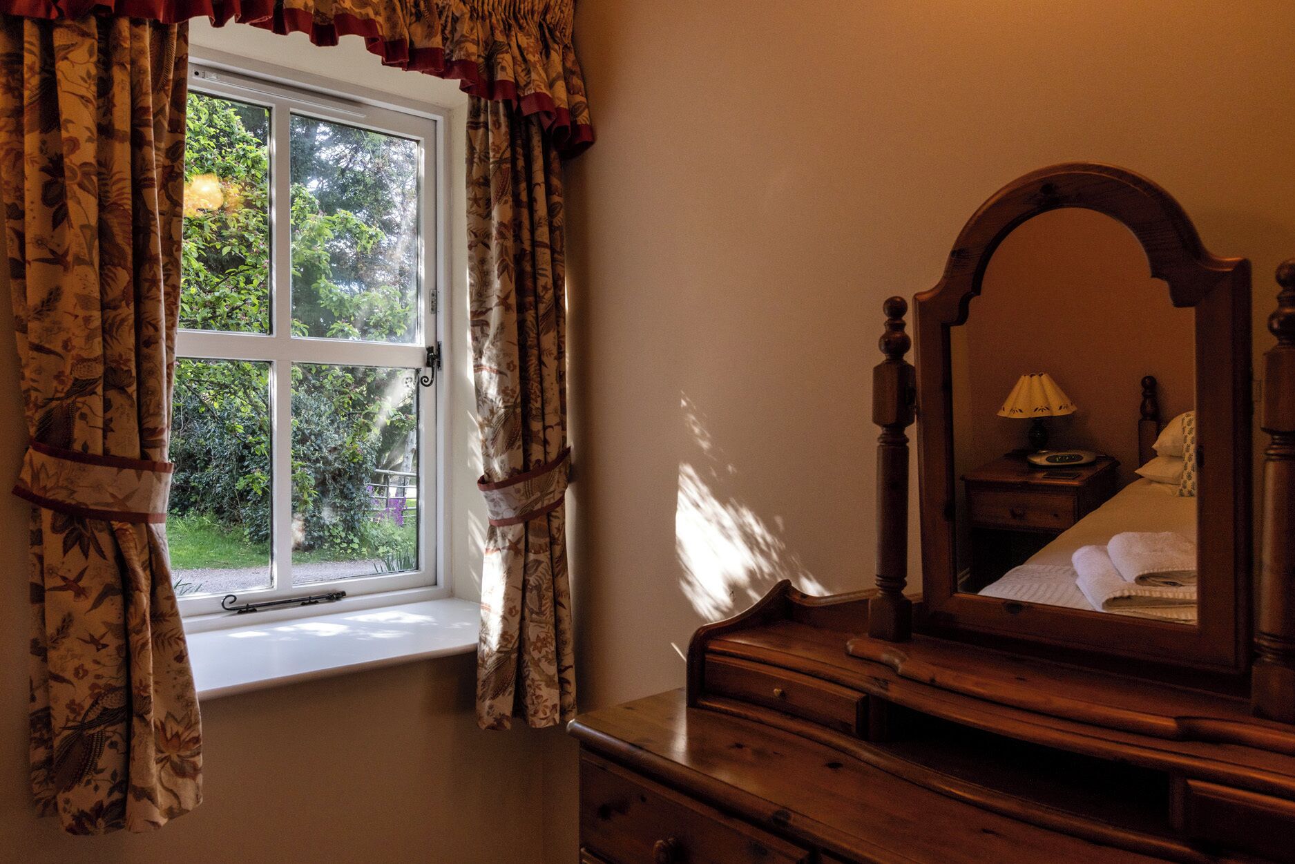 A room with a window, a wooden dresser, and a mirror reflecting a bed with a lamp.