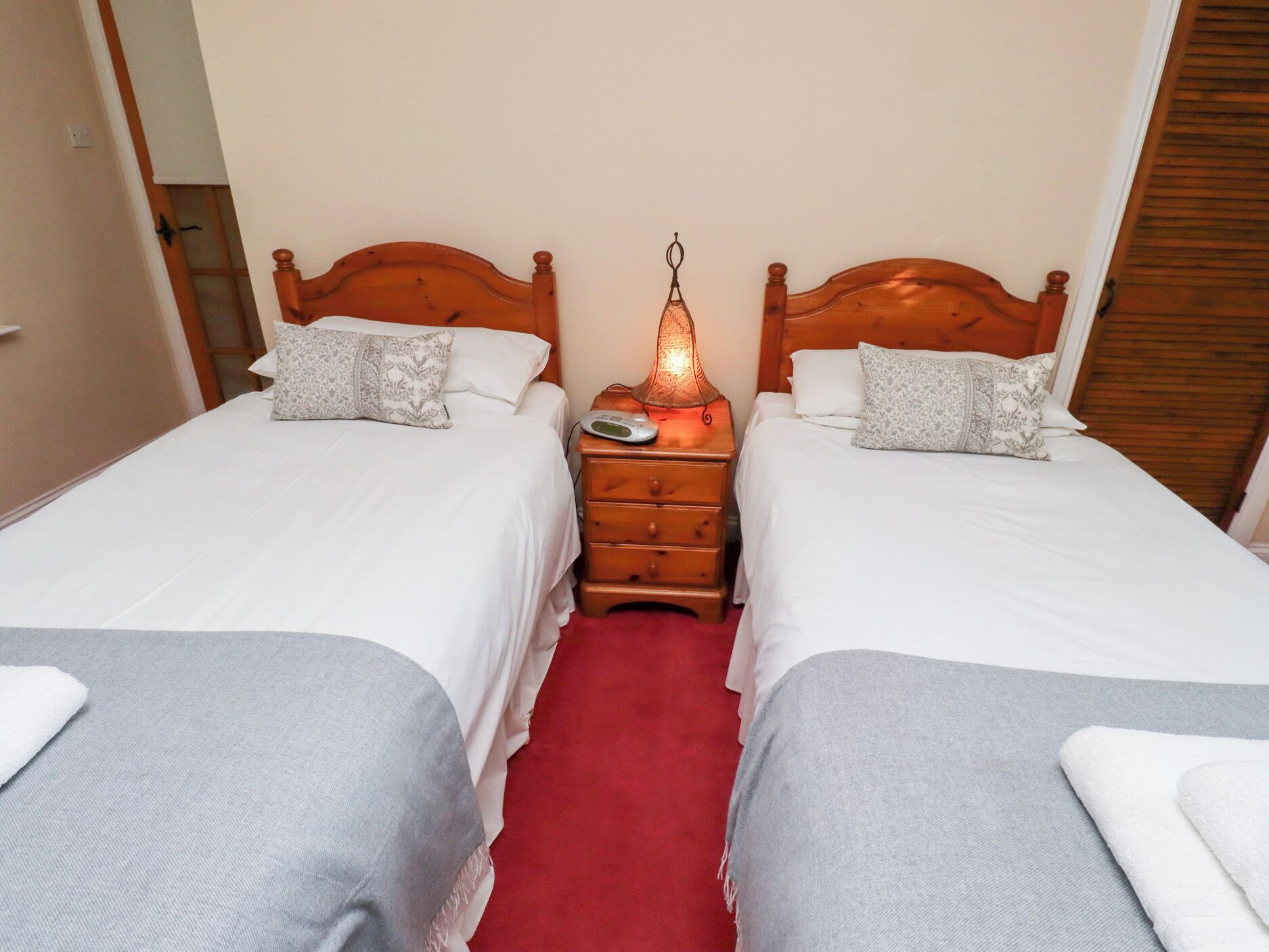 A hotel room with two single beds, a wooden headboard, a nightstand with a lamp, and a red carpet.