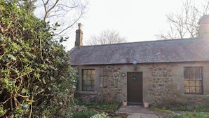 A stone house with a dark door, two windows, and a stone pathway leading to the entrance.