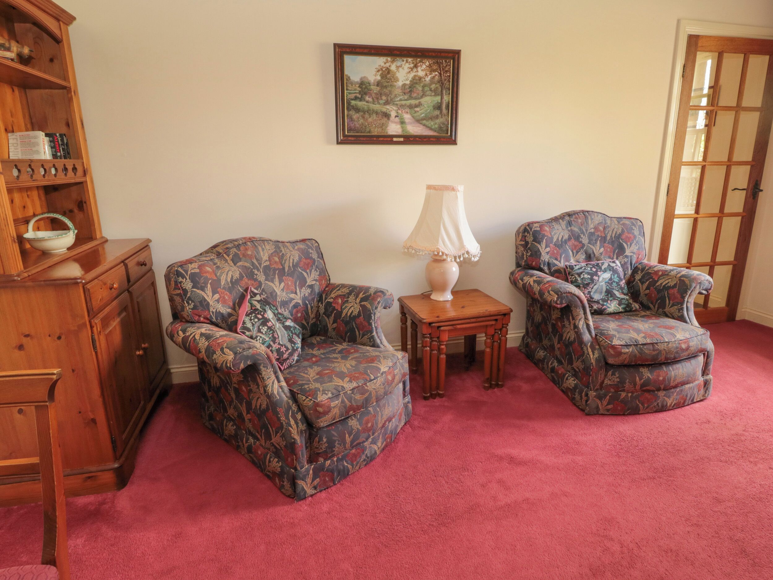 A room with two patterned armchairs, a small wooden table with a lamp, a wooden cabinet, and a framed landscape painting on the wall.