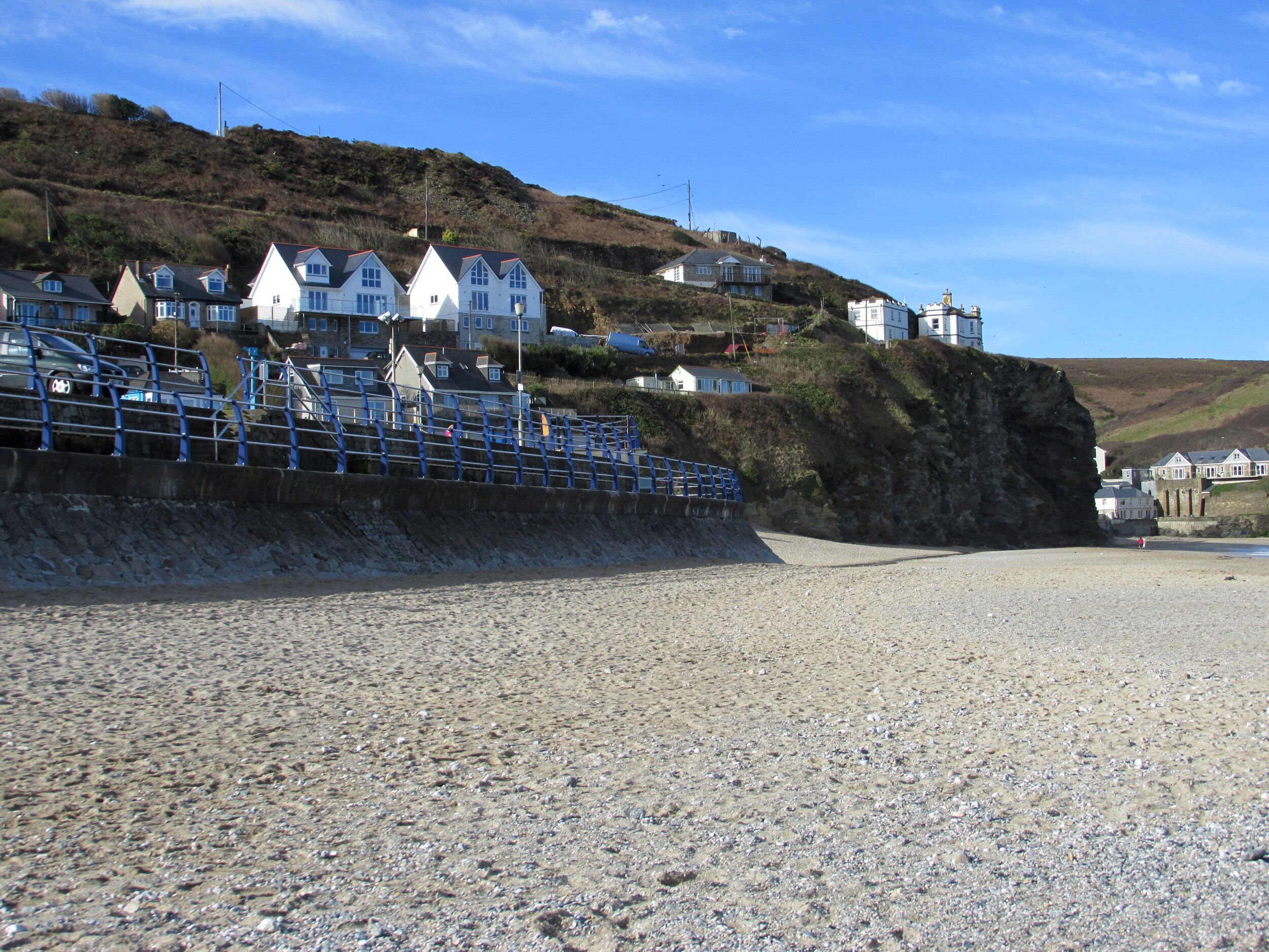 A coastal area with houses on a hillside, a rocky beach, and a blue railing.