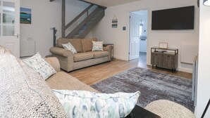 A modern living room with a sofa, a coffee table, a staircase, a TV, and a kitchen in the background. - Beach Cottage (Redruth)