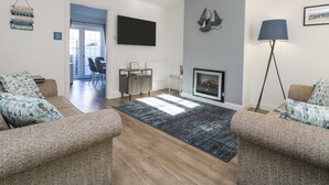 A modern living room with a fireplace, a flat-screen TV, and a dining area visible through an open door. - Beach Cottage (Redruth)
