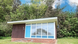 A small, single-story building with a brick base and a brown upper section. It features a large window with white frames and a blue door. - Chalet 126a (Cromer)