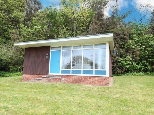A small, single-story building with a brick base and a brown upper section. It features a large window with white frames and a blue door. - Chalet 126a (Cromer)