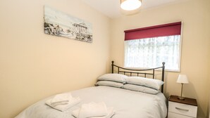A neatly made bed with white linens and pillows, a bedside table with a lamp, a window with a red valance, and a painting on the wall. - Chalet 126a (Cromer)