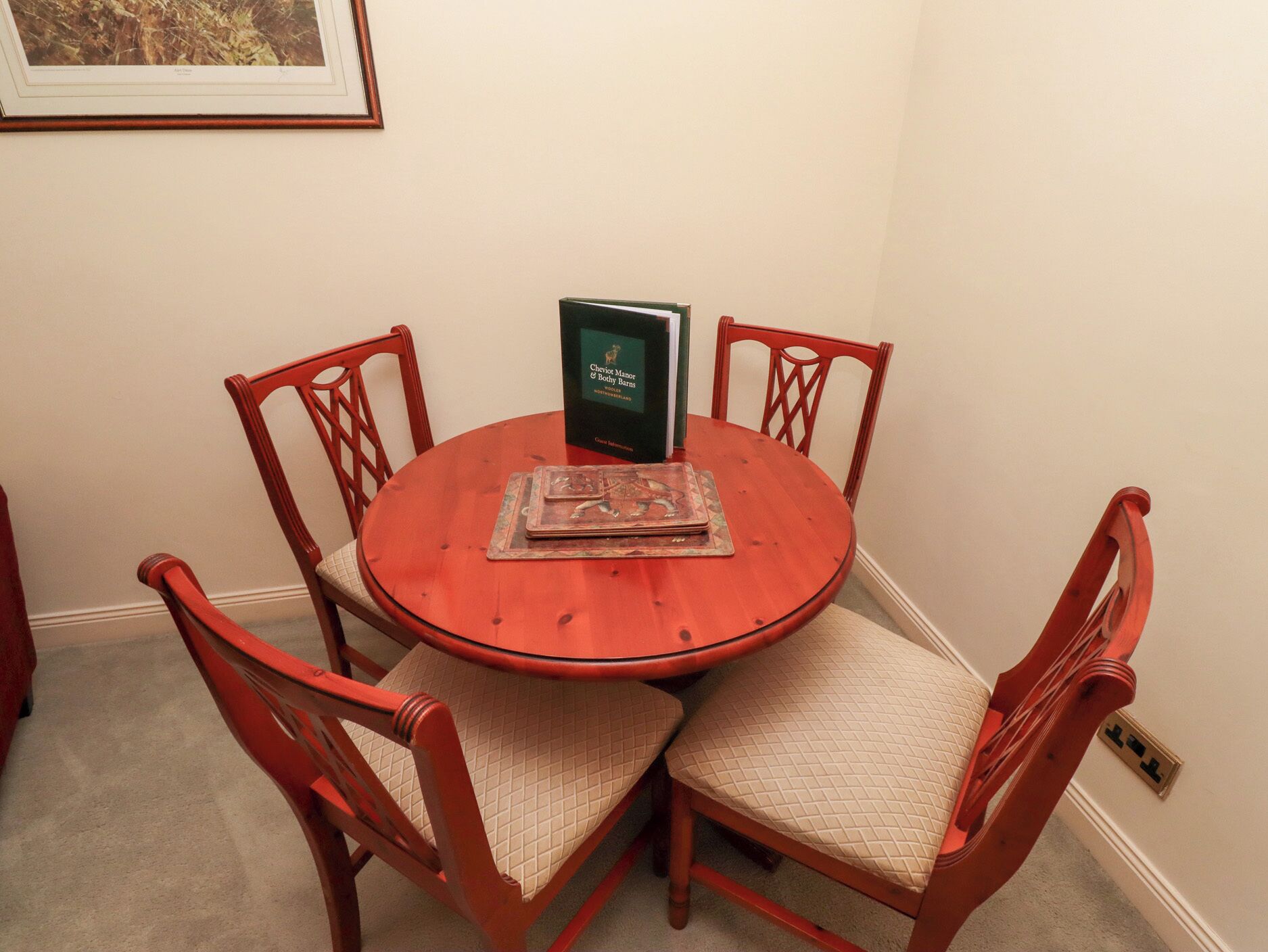 Une table ronde en bois, entourée de quatre chaises, un livre intitulé « Clavijo Masters & Dethy Barns » et un tableau encadré accroché au mur.