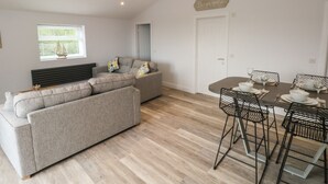 A modern living room with a grey sectional sofa, a dining table with metal chairs, and a window with blinds. - Caban Y Mor (Narberth)