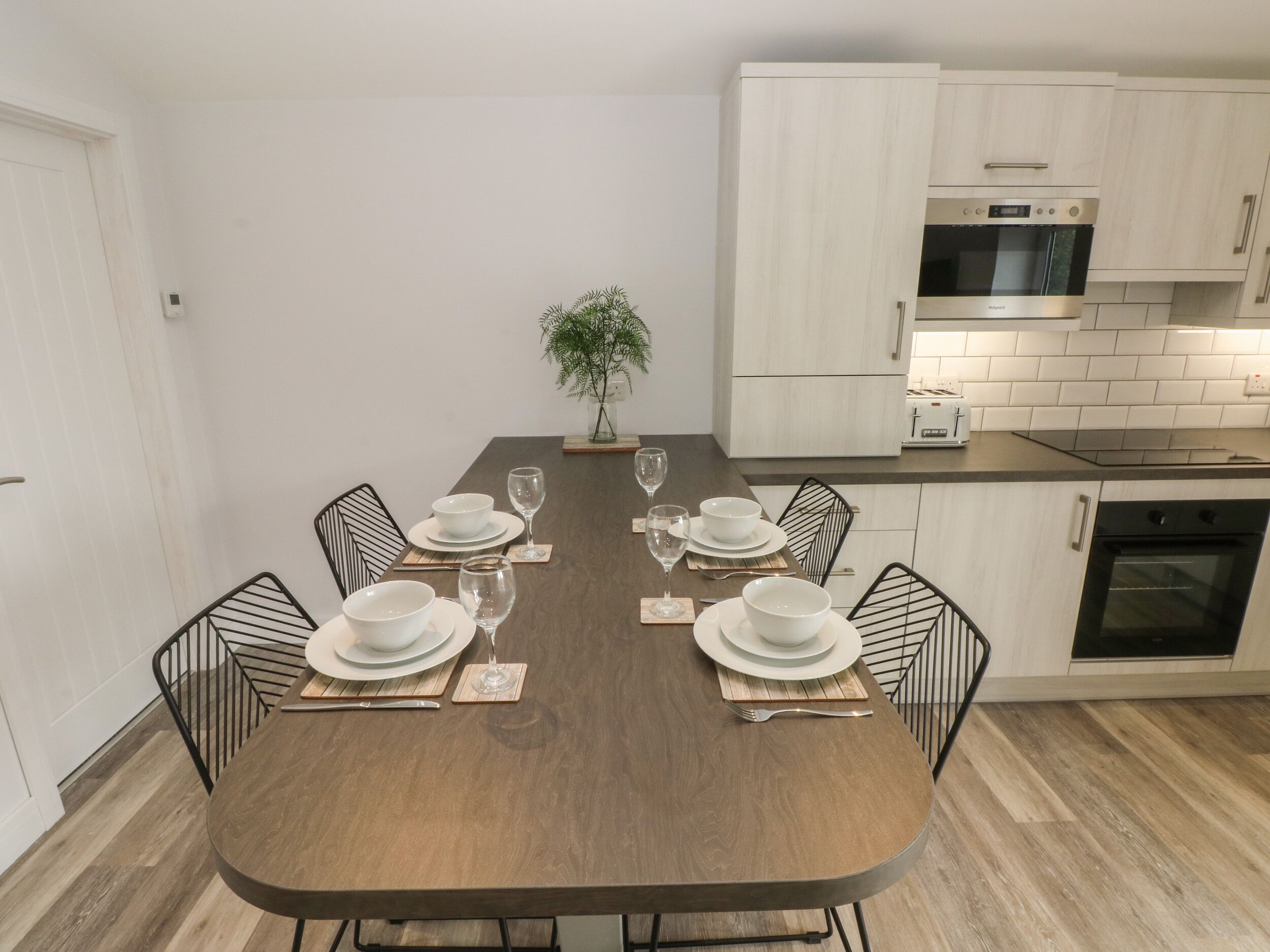 A dining area with a wooden table set for six, surrounded by black chairs. There is a kitchen with white cabinets and a microwave.