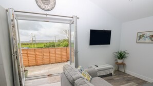 A modern living room with a grey sofa, a small table, a TV, and a sliding glass door leading to a patio. - Caban Y Mor (Narberth)