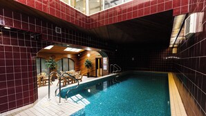 An indoor swimming pool with a tiled floor, a wooden lounge area with chairs, and a small wooden structure with a door.
