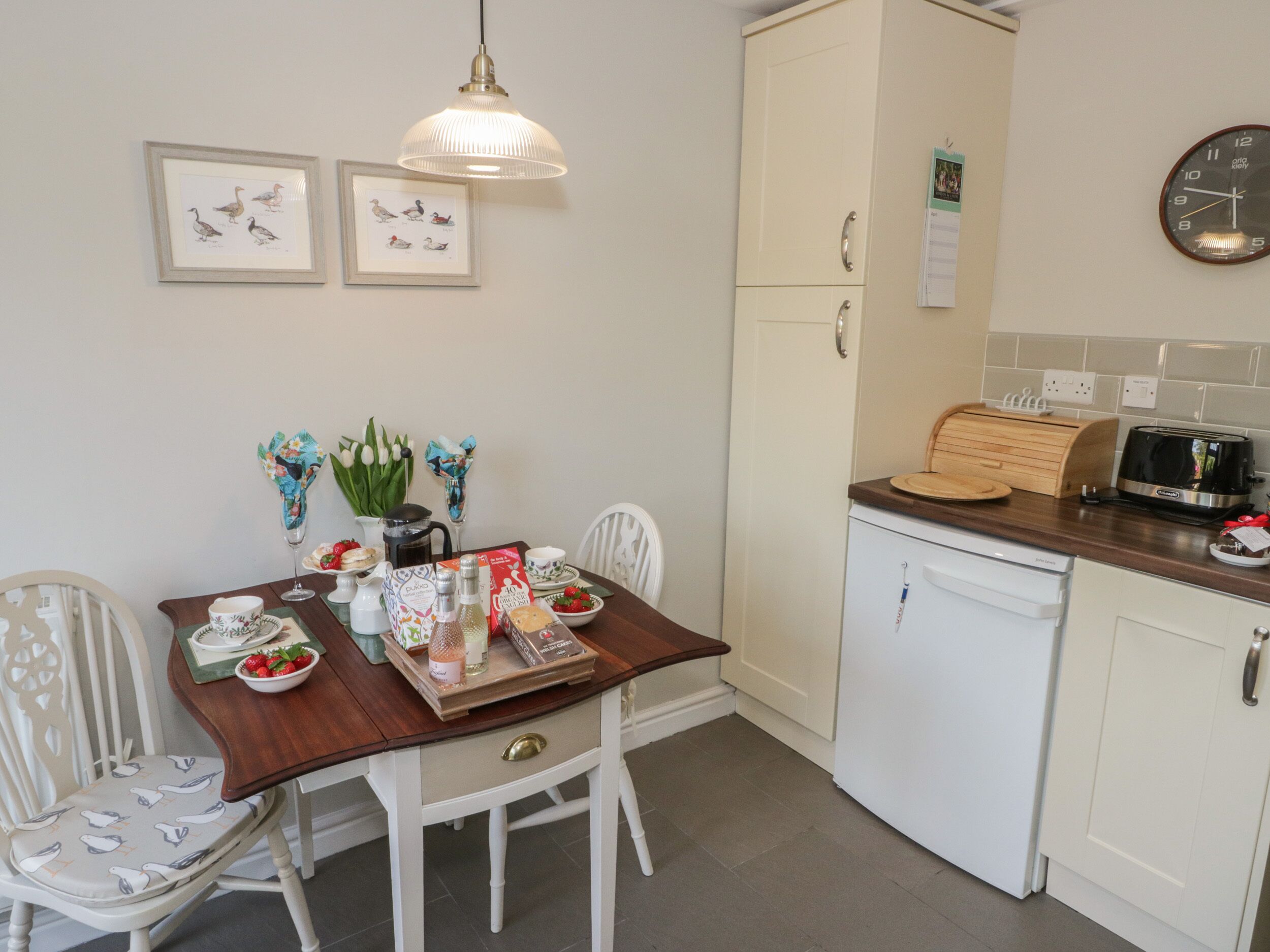 A kitchen with white cabinets, a wooden dining table, white chairs, a coffee maker, a toaster, and framed pictures on the wall.