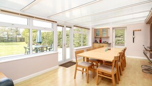 Une pièce spacieuse avec une grande table à manger en bois, plusieurs chaises, un coin cuisine avec des placards et de grandes fenêtres donnant sur l’extérieur.