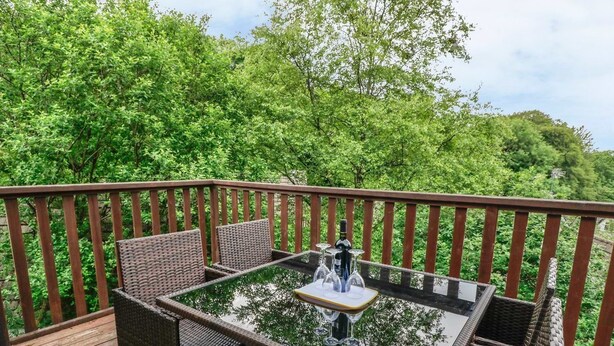 A deck with wicker furniture and a glass table, surrounded by a wooden railing and lush greenery. - 19 Honicombe Park (Gunnislake)