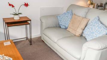 A living room with a light grey sofa, two blue patterned pillows, a wooden side table with a potted plant, and a small table with a plate of food.