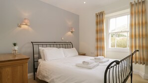 A neatly made bed with white linens and towels, a bedside table with a lamp, and a window with curtains. - Glen Cottage (Penrith)